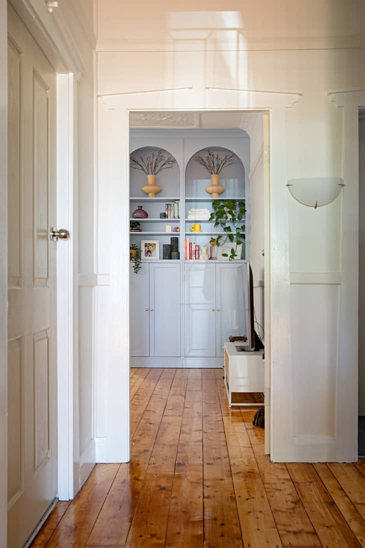 A hallway to a light blue built-in bookshelf filled with decorative pieces and cabinets in a white living room.