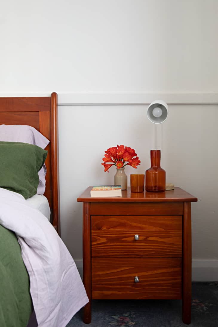 A wooden two-drawer nightstand with books, a lamp, and glass vases.