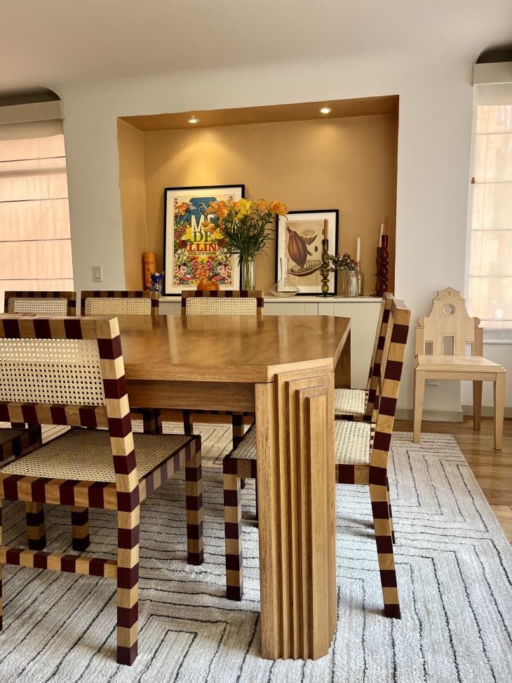 White dining room with one tan accent wall, "striped" wood and rattan dining chairs, and framed posters and flowers on shelf