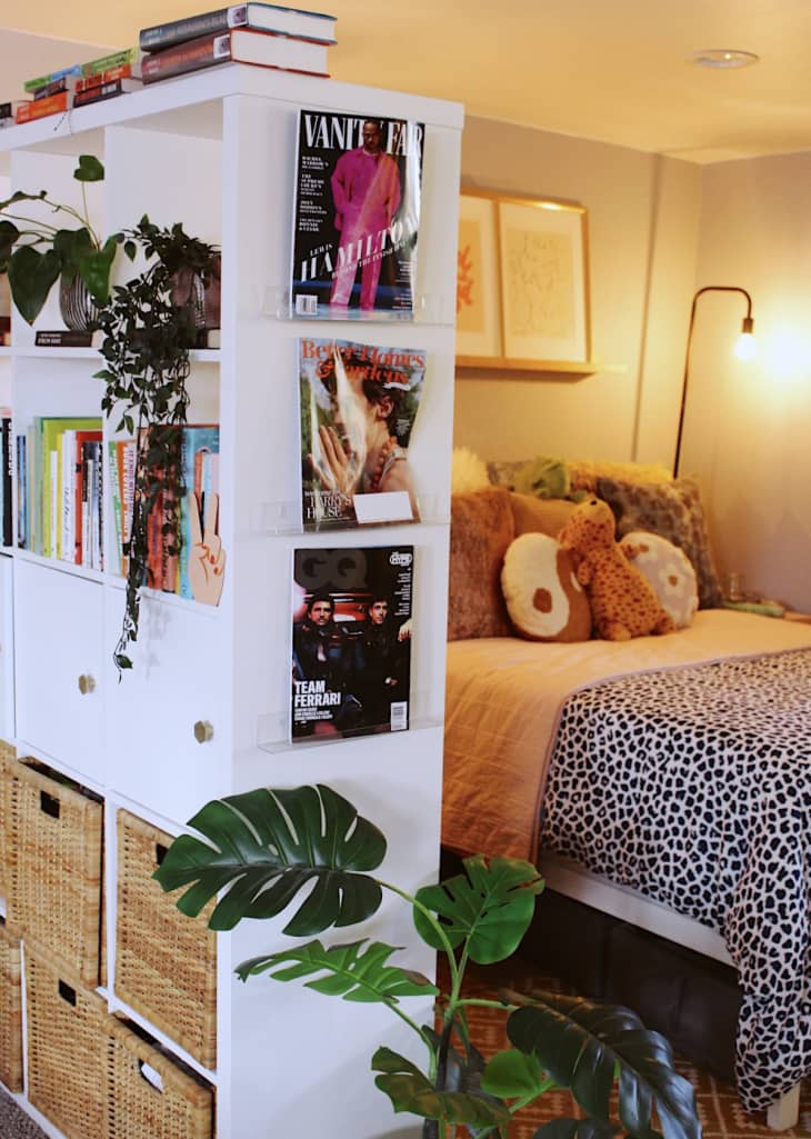 Studio apartment with bedroom area divided from living space by white shelves with books, plants, and basket bins