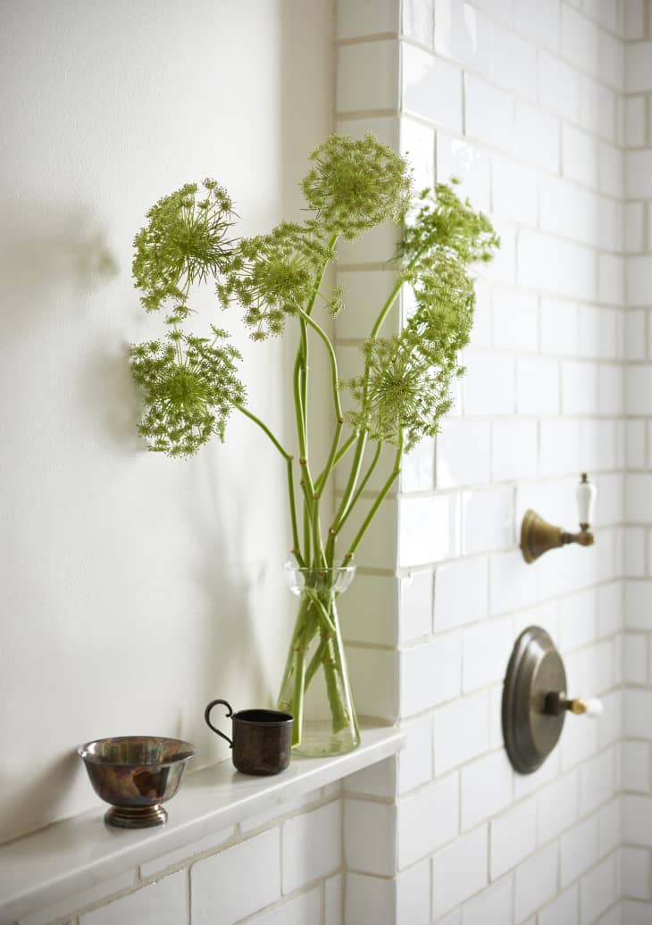 A vase holds plants on a shelf in a white tile bathroom.