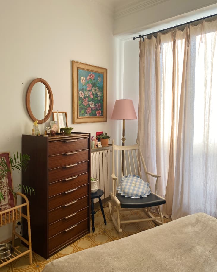 corner of white bedroom with tall narrow wood dresser, rocking chair, and large windows with sheer curtains