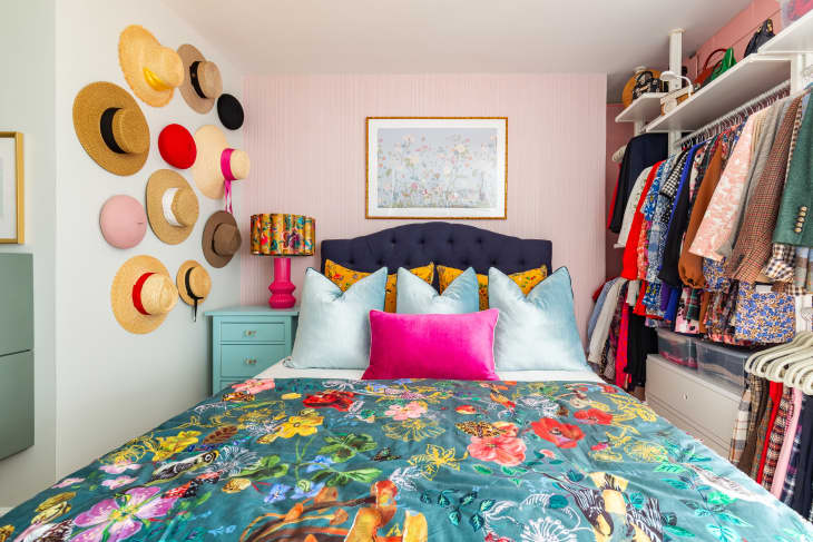 A bed with colorful sheets and pillows flanked by sunhats and clothing hung in a open-face closet.