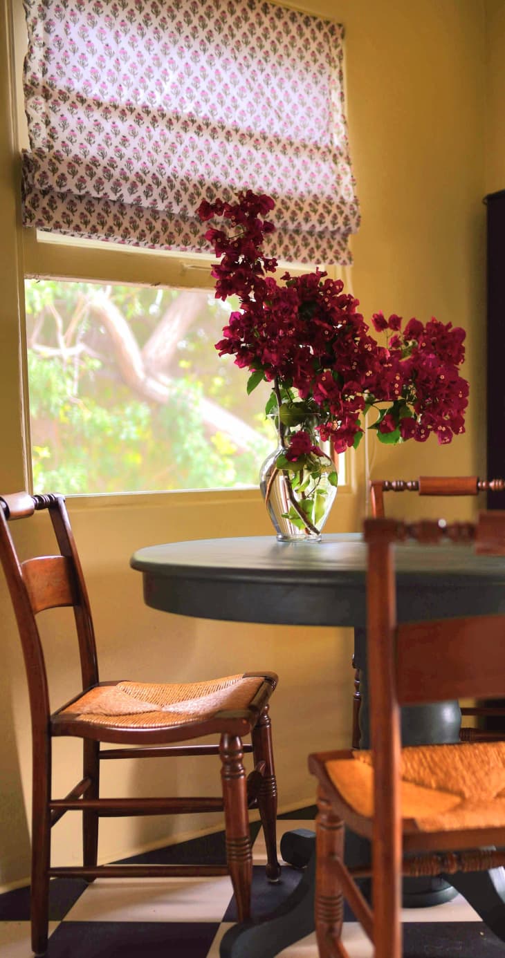 Yellow dining room with round table and wood/rattan chairs, patterned window shade, and vase with bougainvillea