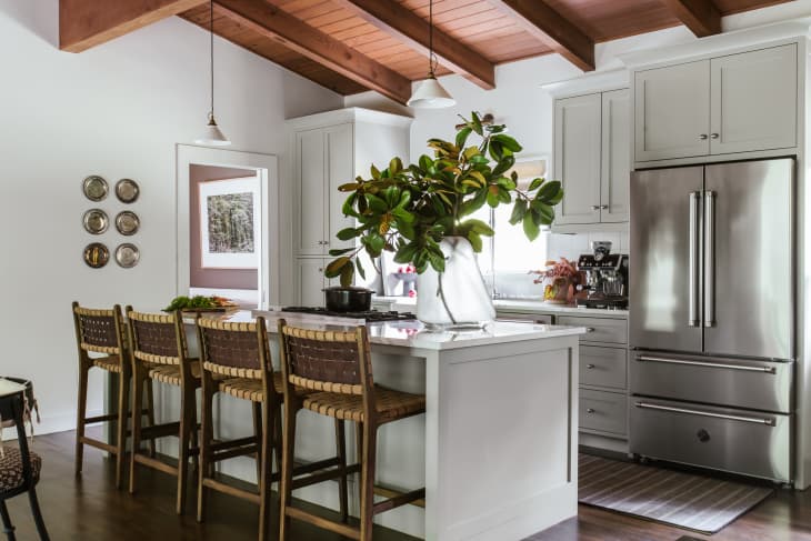 Large kitchen island, white marble countertop with gray veining, large woven back bar stool, large plant on countertop, red wood ceiling beams