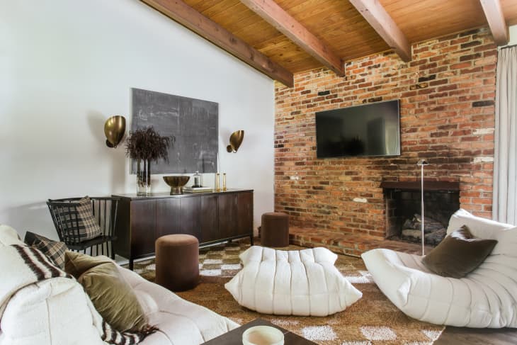 Red wood ceiling with large beams, brick accent wall with tv, brown and cream checkered rug, soft white modern shaped couches