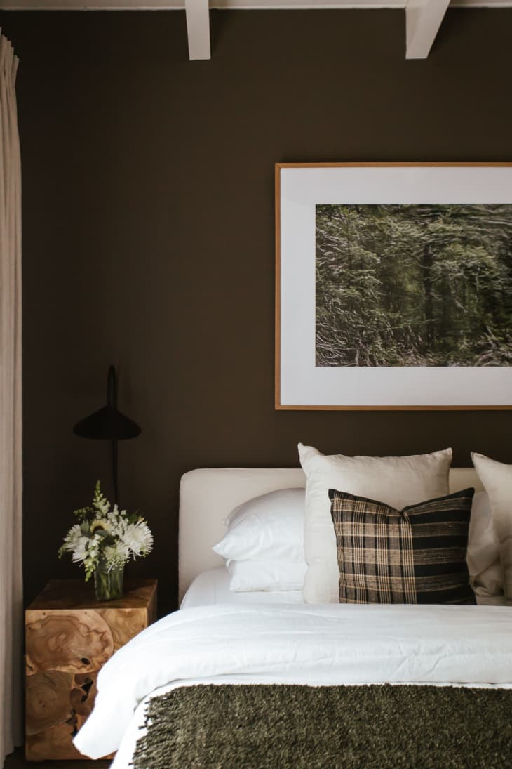 Dark green walls with white fabric headboard, white linens on bed, dark green throw blanket, landscape forest art on wall