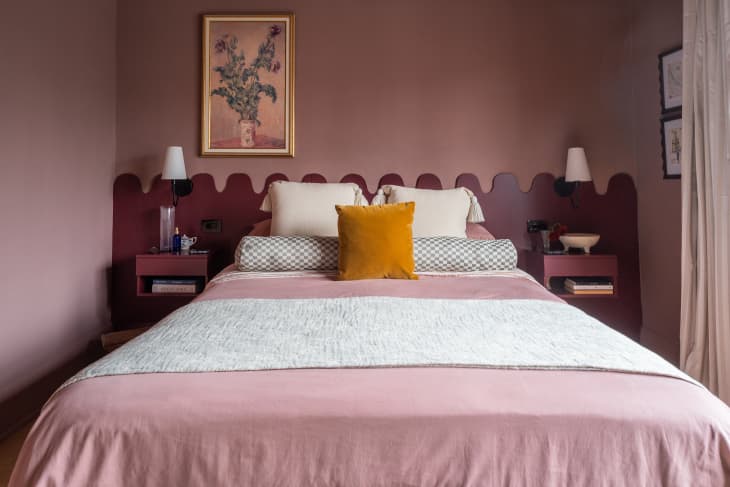 A bed with a burgundy built-in head board and an orange decorative pillow.