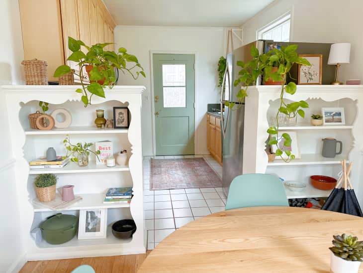 Vintage built in book shelves dividing kitchen from dining room, sage green door matching dining chairs, light wood cabinets, white tile floor.