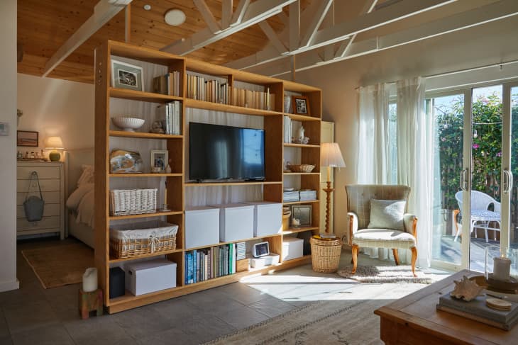 A large media bookcase with TV books and decorative items divides the bedroom from the living room area.