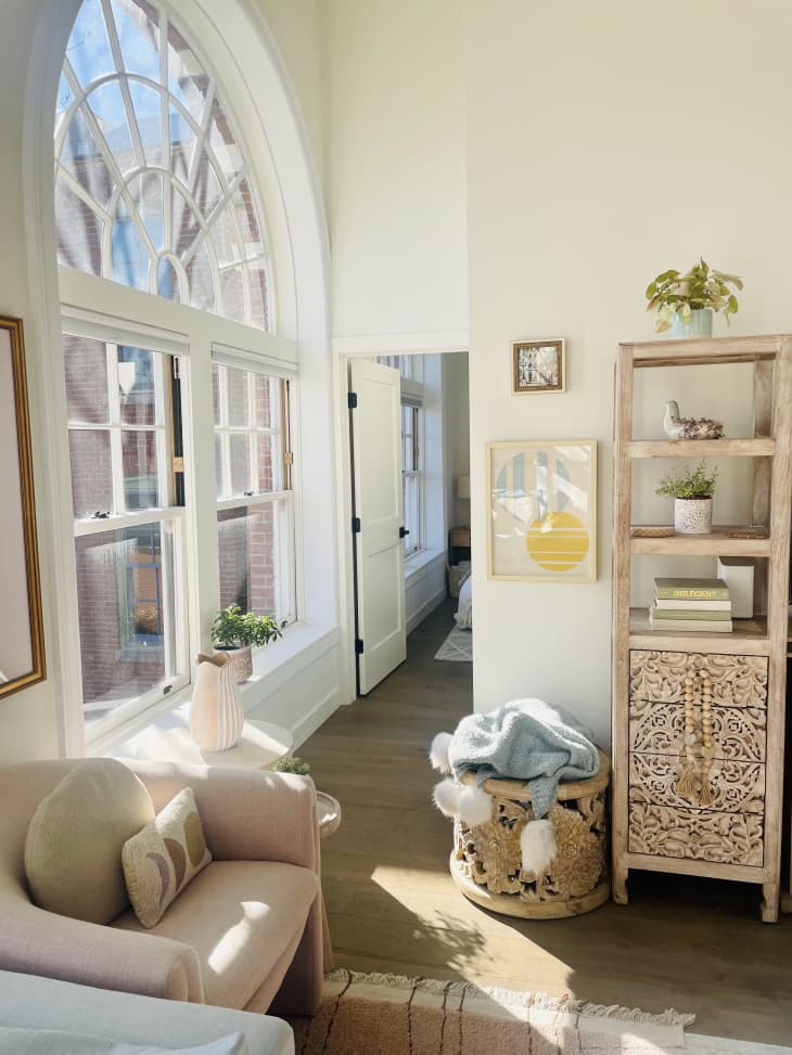 Arched picture window with lots of natural sunlight, pink modern fabric arm chair, tall vertical wood cabinet with shelving and drawers and etchings in wood, blanket basket on floor