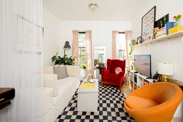 Cozy living room featuring a white sofa, red armchair, orange accent chair, and a checkered rug. Decor includes plants and shelves.