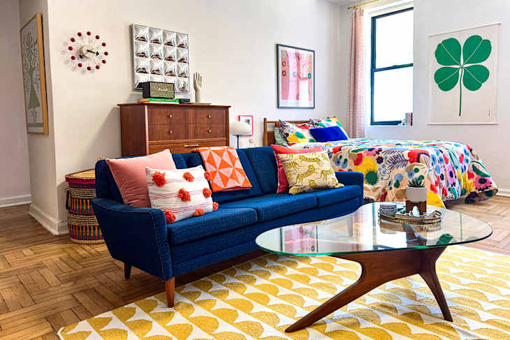 Colorful living space featuring a blue sofa with patterned pillows, a glass coffee table, and a floral bedspread.