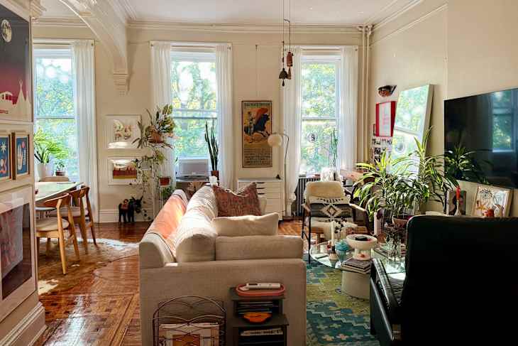 Bright living room with a beige sofa, plants, artwork, and a glass coffee table on a colorful rug.
