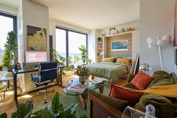 Cozy studio apartment with a bed, desk, plants, and colorful decor, featuring large windows and a view.