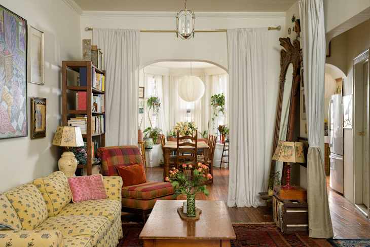 Cozy living room with a yellow floral sofa, plaid chair, wooden coffee table, and a dining area with plants and a lantern.