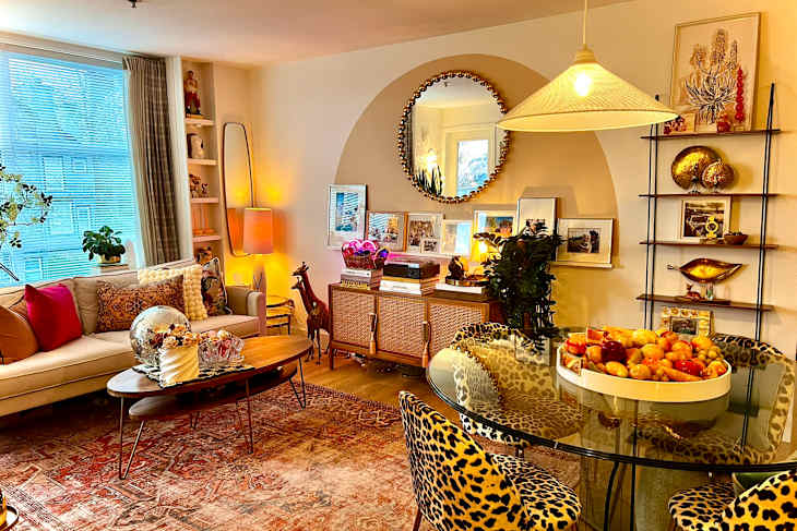 Cozy living room with a beige sofa, leopard print chairs, a round glass dining table with a fruit centerpiece, and decorative shelves.