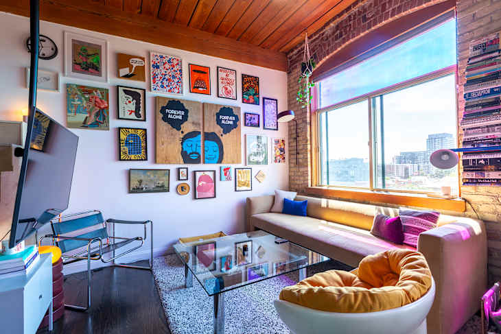 Cozy living room with a large window, colorful artwork on the wall, a glass coffee table, and a beige sofa with decorative pillows.