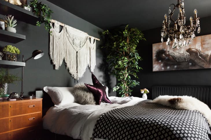 A black bedroom with a chandelier, black and white bedding and white macrame on the wall.