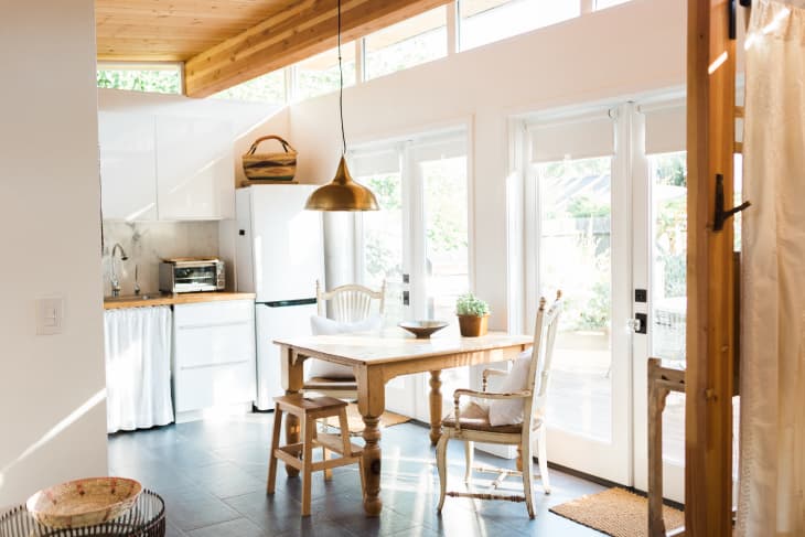 Bright kitchen with wooden table, chairs, pendant light, and potted plant near large windows and white cabinets.