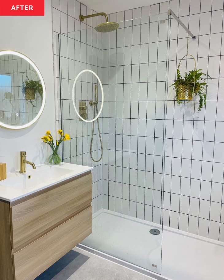White vertical subway tile in newly renovated bathroom with glass shower door and white neon light around rounded mirror mounted above wooden bathroom vanity.