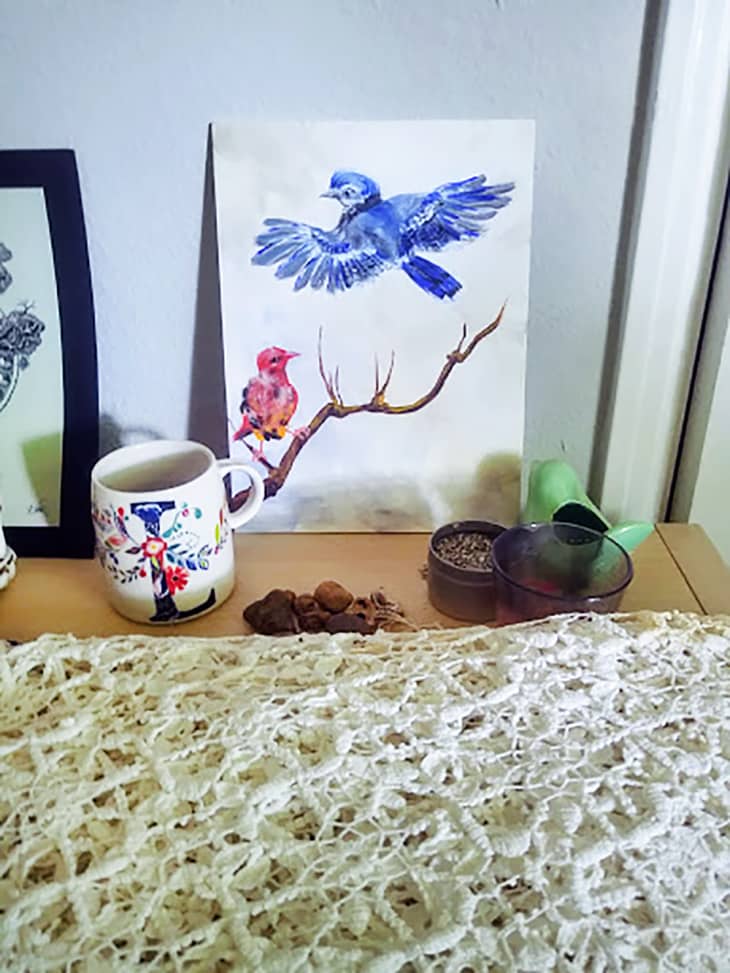 A tablecloth sits folded on a wooden shelf, surrounded by a mug, a painting, and a few other trinkets
