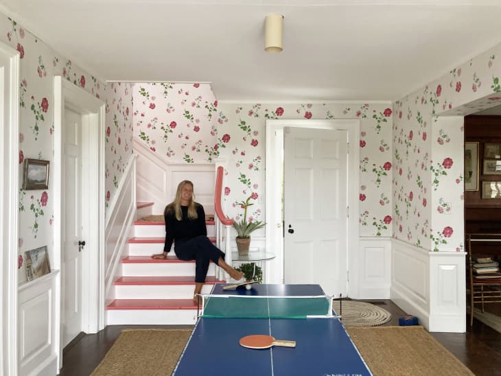 A woman sits on the stairs in the floral-wallpapered entryway of a beach house with a ping pong table