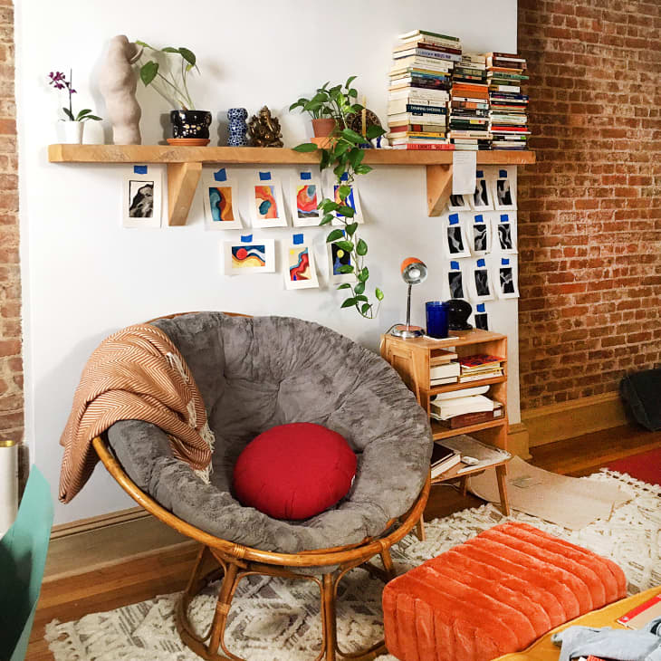 A plush gray papasan chair on a wooden frame, inside an artful bohemian apartment with a brick wall, stacks of books, and small pieces of artwork taped to the wall