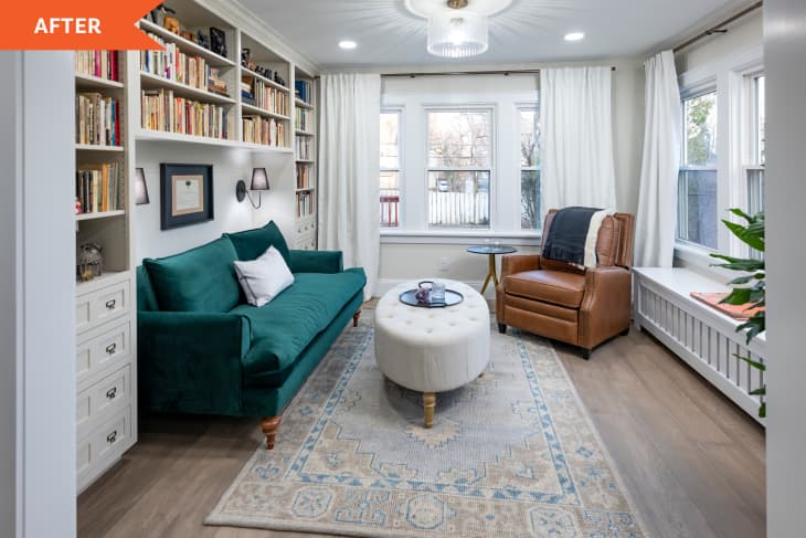 "after" photo of a living room with white walls, white ceiling-t-floor curtains, a dark teal sofa, brown leather armchair. There are built-in bookshelves full of books. A white large ottoman doubles as a coffee table in the center of the room. Vintage-look patterned rug is centered on a wood or laminate floor