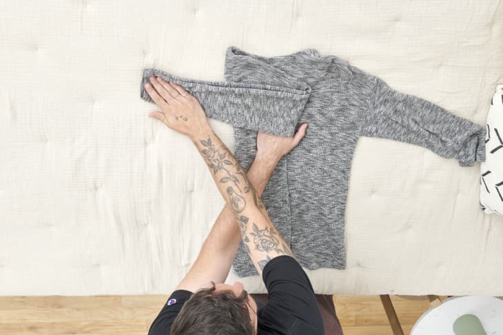Overhead view of man folding the arms of a grey sweater on a beige duvet.