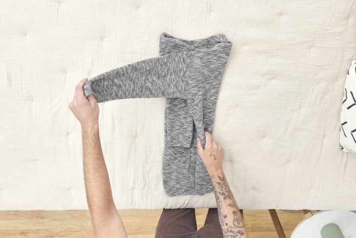 Overhead view of a man folding the right side of a grey sweater into the center, on a beige duvet.