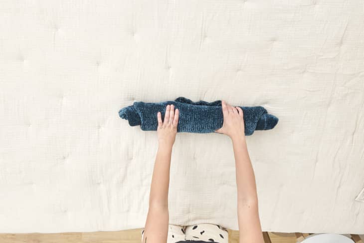 Overhead view of woman rolling a blue sweater on a beige duvet.