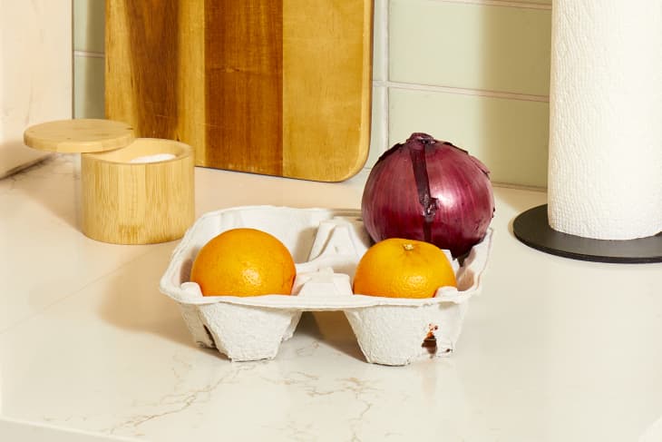 Head on shot of a cardboard drink holder sitting on a white marble counter, holding a red onion and two oranges in it.