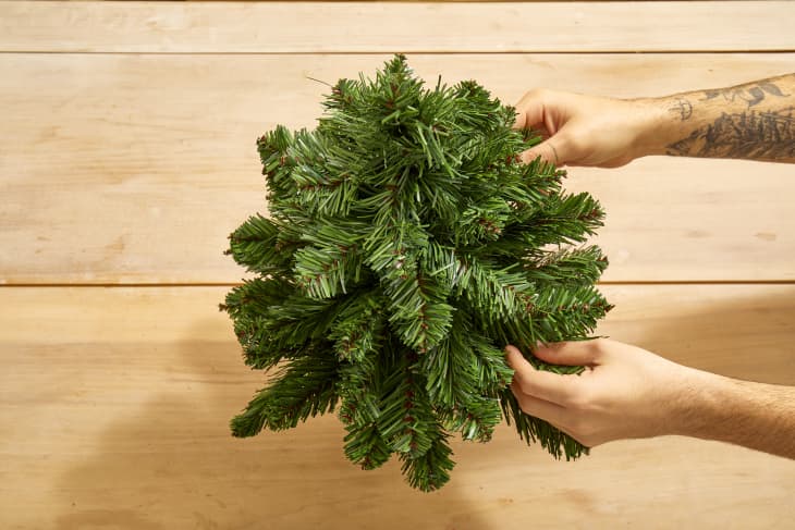 Overhead shot of two hands reaching in from the right, to adjust and fluff a small faux christmas tree on a light wood surface.