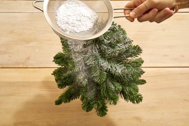 Overhead shot of a small christmas tree being dusted with fake snow on a light wood surface.