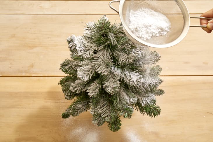Overhead shot of a small christmas tree being dusted with fake snow on a light wood surface.
