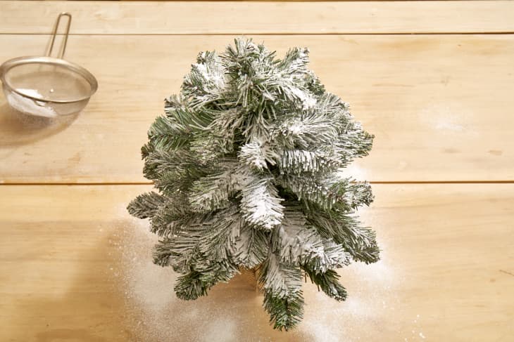 Overhead shot of a small christmas tree dusted with fake snow on a light wood surface.