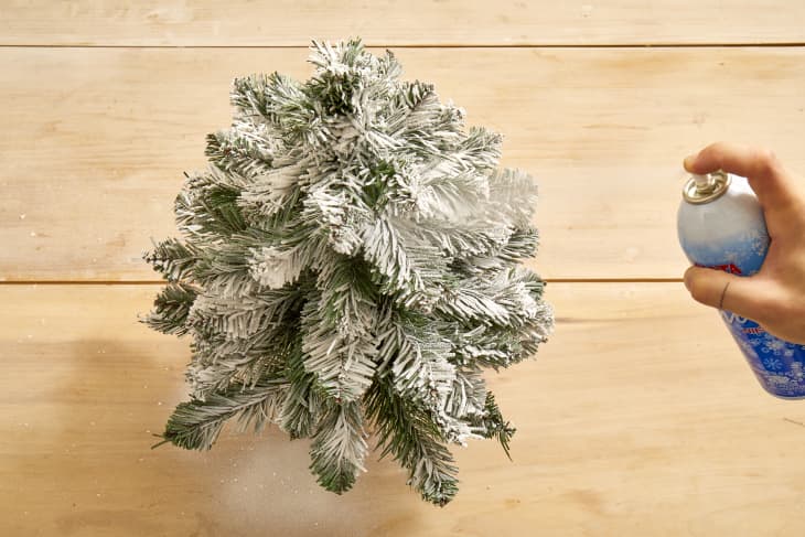 Overhead shot of a small christmas tree being sprayed with fake snow on a light wood surface.