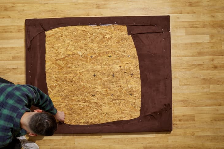 Overhead shot of a man wearing a blue and green plaid shirt while stapling dark brown fabric to a large piece of ply wood on a light wood floor.