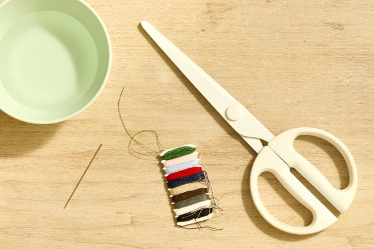 Overhead shot of a pair of white scissors, a bundle of thread, a needle and a small bowl of water.