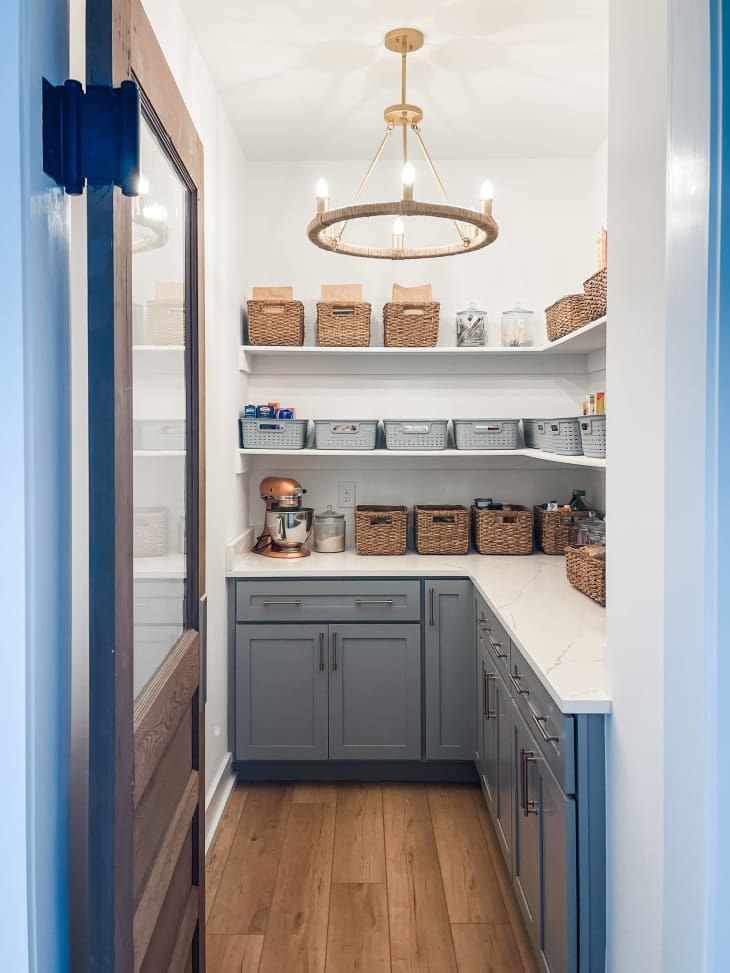 light blue lower cabinets with white counter top and exposed floating shelves with organization baskets and a small round chandelier, blue door with window on top