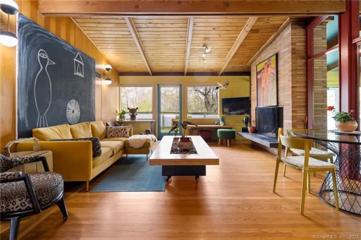 large open living room with wood walls and ceiling, gold sofa, large coffee table, large chalkboard painting behind sofa. Wire/metal dining table on right side of screen with yellow chairs