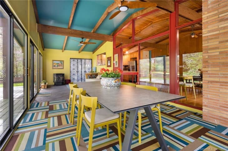 Dining room with colorful walls and ceiling, yellow dining chairs, colorful area rug with stripes/squares, one wood wall, exposed wood ceiling beams, large windows on both sides of room