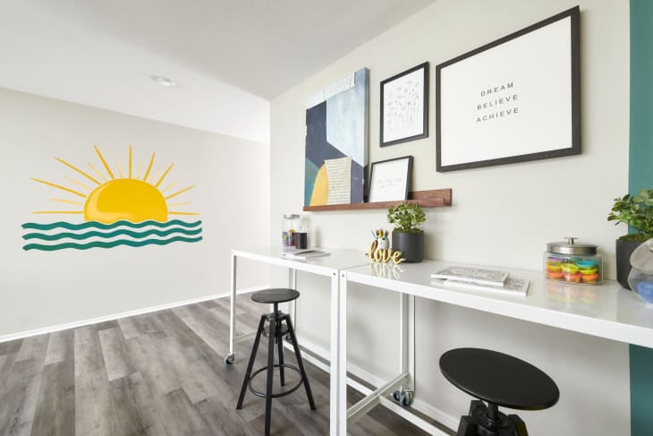 Home office space with white narrow desk lining length of wall, black stools, floating shelf with artwork, sunshine/water mural on white wall