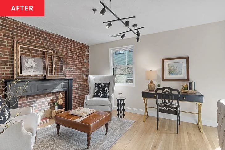 living room after home staging: white walls, one exposed brick wall, 2 seating areas, one with 2 tall white armchairs and leather ottoman/table, faux fireplace, one with white sofa, pale gray area rug, media console, glass coffee table, leather armchair