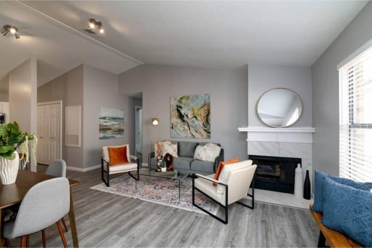 Matching modern white arm chairs, gray couch, orange pillows, gray wood floors, fireplace in corner, coral colorful rug