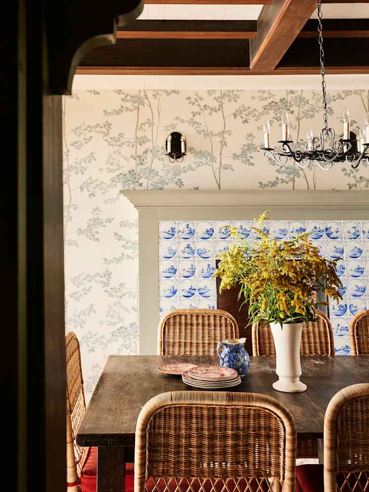 Dining room featuring a wooden table, woven chairs, floral wallpaper, and a vase with yellow flowers.