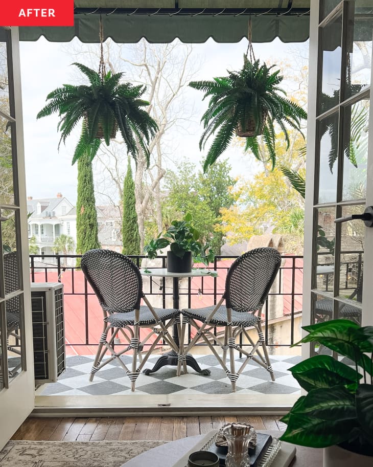 View of small terrace from living room after makeover. Black and white checkered painted floor, small round table with 2 woven chairs, plants in corners, 2 large ferns hanging