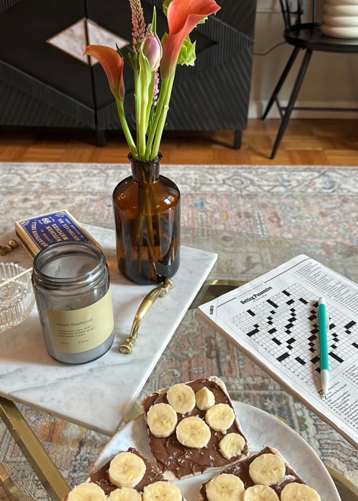 H&amp;M scented jar candle sitting on small table next to vase of flowers, some banana and nutella toast, and a crossword puzzle
