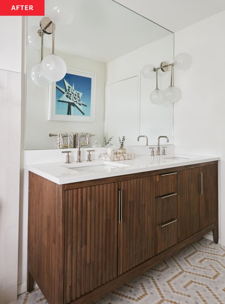 Bathroom after renovation/makeover: modern wood sink cabinet, white countertops, dual sinks, 2 globe sconces, silver hardware, mosaic pattern tile floor, white tile and white walls